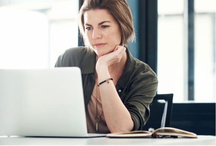 Woman sitting behind a laptop at a desk resting her head on her hand.