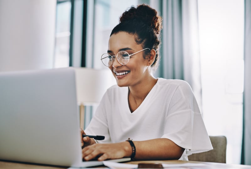 Smiling woman sat behind a laptop at a desk in a brightly lit room.